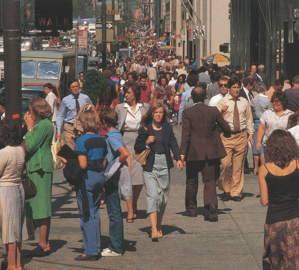 A busy city sidewalk filled with people walking in various directions. Most are dressed in business or casual attire, and the street is crowded with activity on a sunny day.