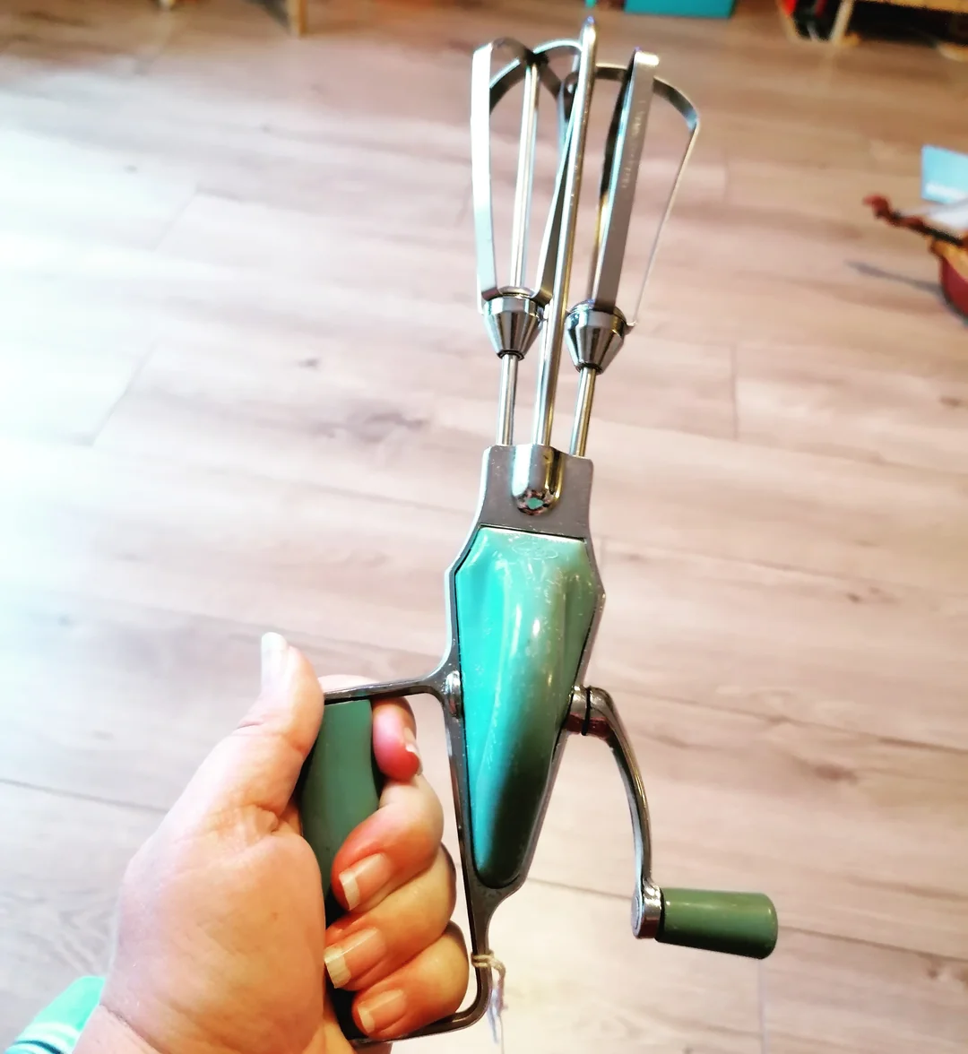 A hand holds a vintage green manual egg beater with two metal beaters and a hand crank, against a wooden floor background.