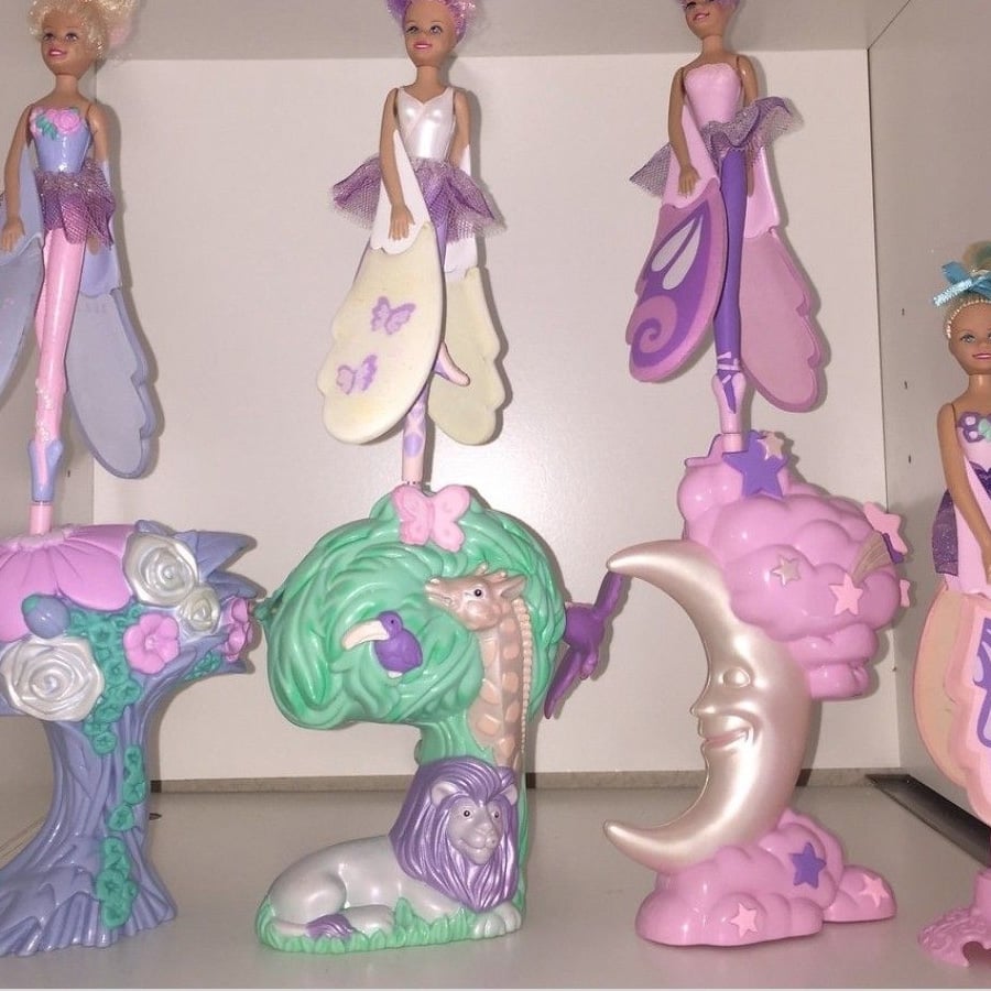 Four fairy dolls with butterfly wings stand on pastel-colored stands shaped like a tree with a lion, a crescent moon with clouds, and flowers, all set on a white shelf.