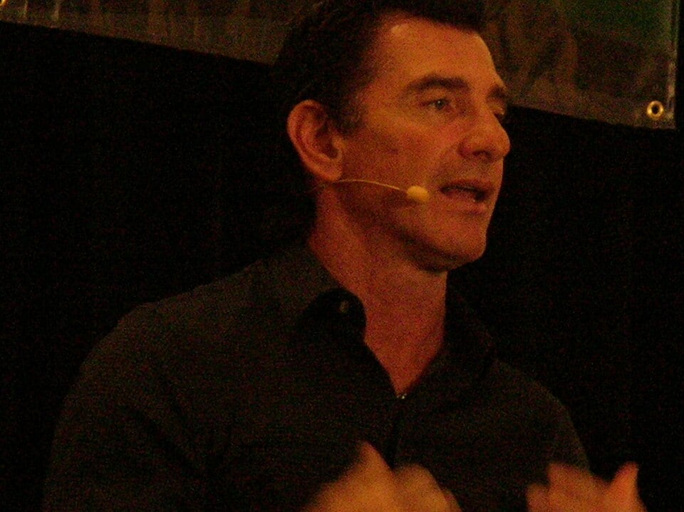 A man with short dark hair, wearing a dark shirt and a headset microphone, speaks in front of a dark background.