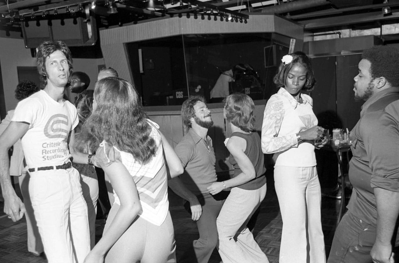 A group of people dance and socialize in a lively nightclub, wearing 1970s-style clothing. The scene features energetic movement, retro fashion, and a background with bar and stage elements.