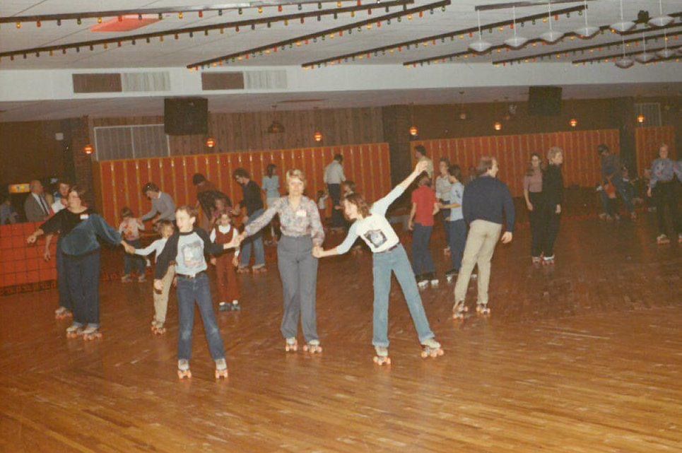 People roller skating in a retro indoor rink with wood floors, orange lockers, and groups holding hands. The scene is lively, with skaters of various ages enjoying the activity under ceiling lights.