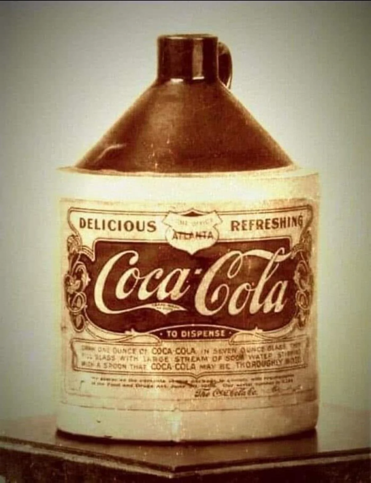 A vintage jug labeled “Coca-Cola” with ornate text, reading “Delicious Refreshing,” and dispensing instructions, evoking an old-fashioned soda fountain style.