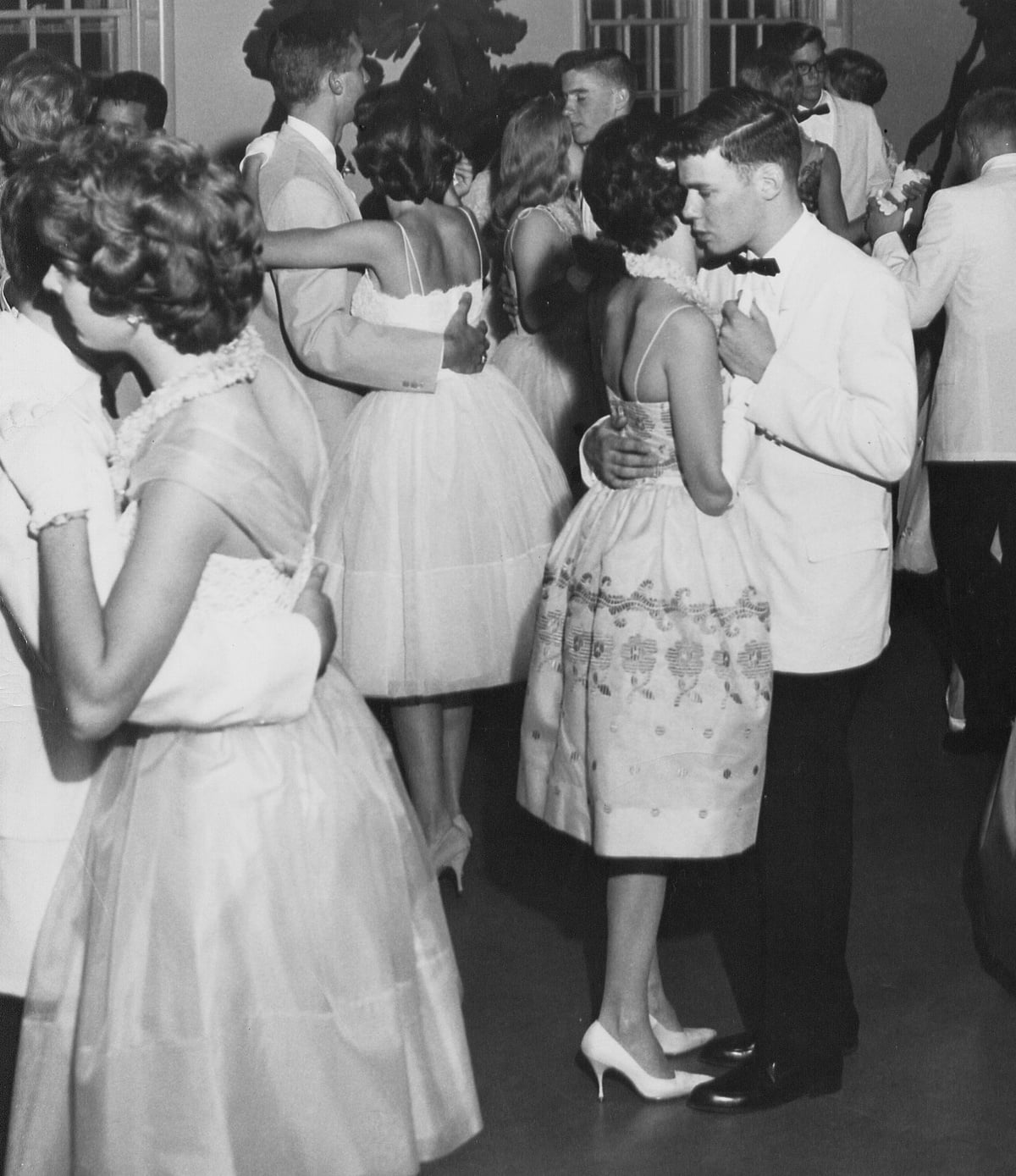 Teen couples in formal attire slow dance at a 1950s or 1960s event. The girls wear dresses and heels, the boys wear suits or tuxedos. The atmosphere is elegant and classic.