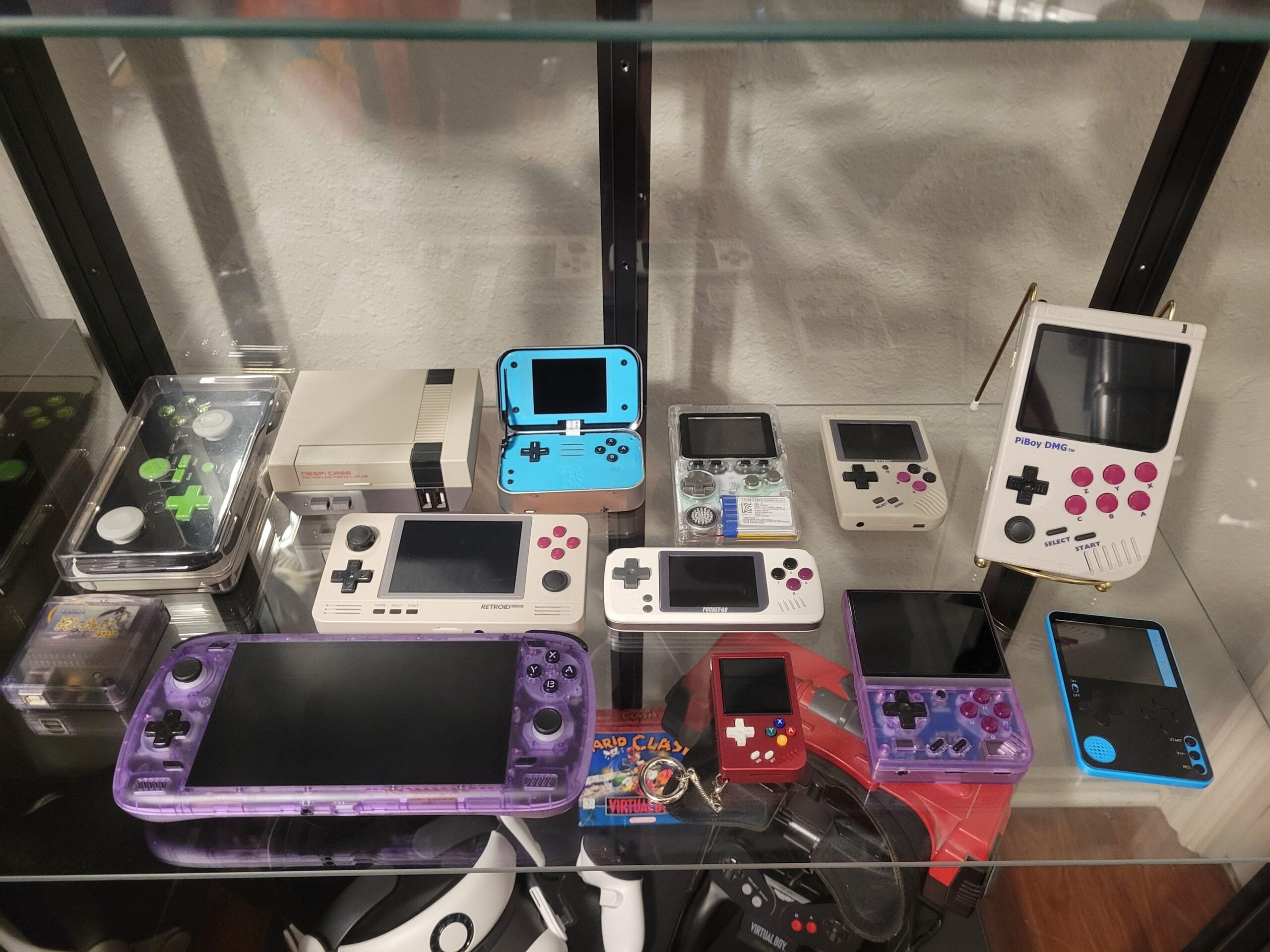 A display case with various retro handheld gaming consoles, including Game Boy-style devices, a mini NES, and other vintage and modern handheld systems in different colors and styles.
