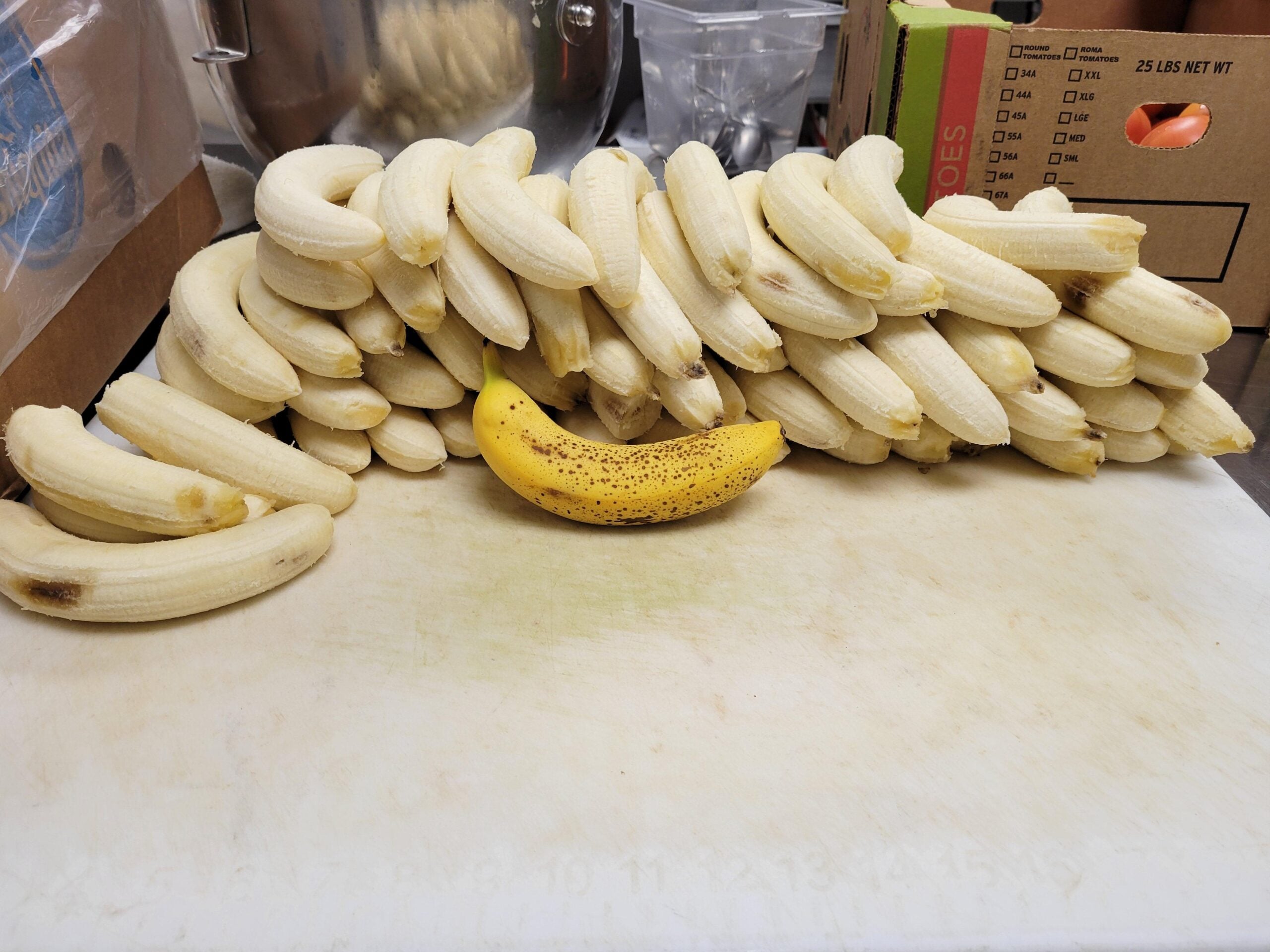 A single unpeeled banana sits in front of a stack of peeled bananas on a white cutting board, surrounded by cardboard boxes and kitchen equipment.