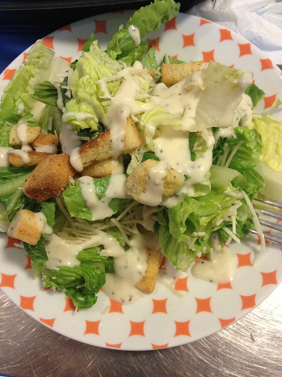 A plate of Caesar salad with romaine lettuce, croutons, shredded cheese, and creamy dressing, served on a patterned dish with a fork visible on the right.