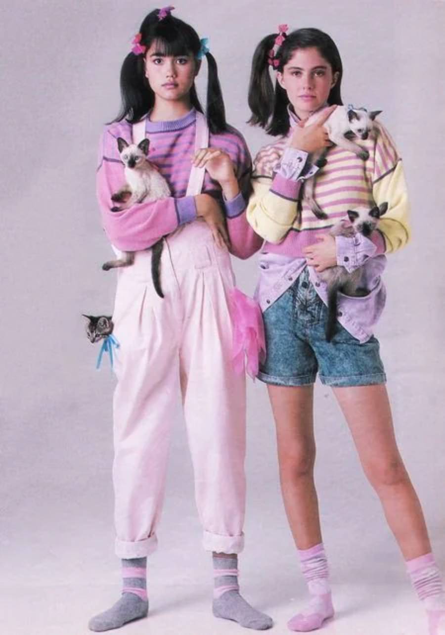 Two young women in pastel 80s outfits holding Siamese cats. One wears pink overalls, the other wears denim shorts. Both have pigtails, striped sweaters, and socks. Several cats are tucked into their clothes.