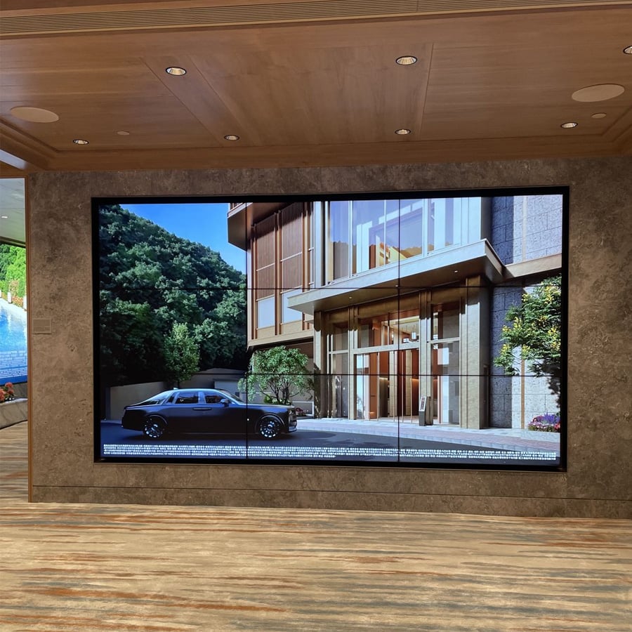 Large digital screen displays an image of a modern building entrance with a parked black car and trees, mounted on a wall in a room with wooden ceiling and patterned carpet.