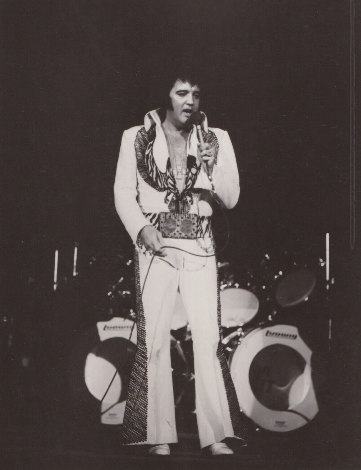 A man in a white jumpsuit with a wide belt sings into a microphone on stage, standing in front of a drum set.