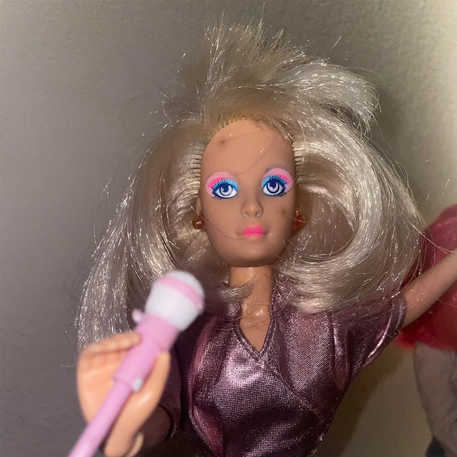 A blonde fashion doll with bright blue eyes and pink eyeshadow holds a pink microphone, wearing a shiny purple outfit.