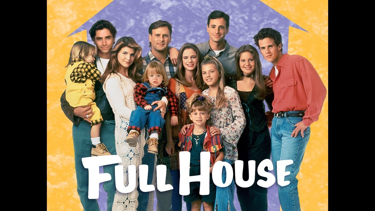 The cast of “Full House” stands together, smiling, in front of a yellow and purple house-shaped backdrop. The show’s title appears in bold white letters at the bottom.