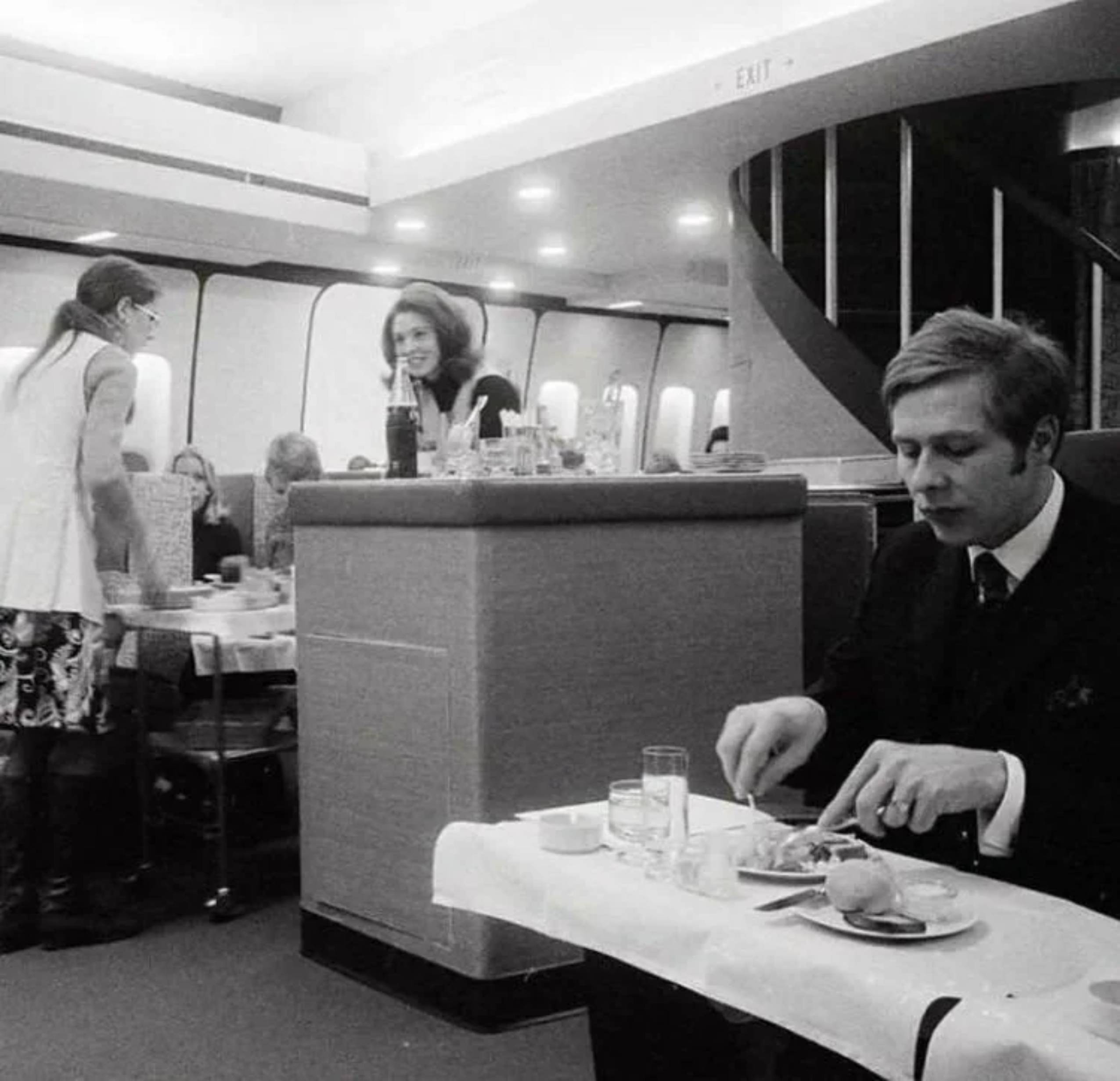 A man in a suit eats at a small table on a plane. Nearby, a woman stands by a counter with drinks. Another woman sits, and a spiral staircase is visible in the background. The image is in black and white.