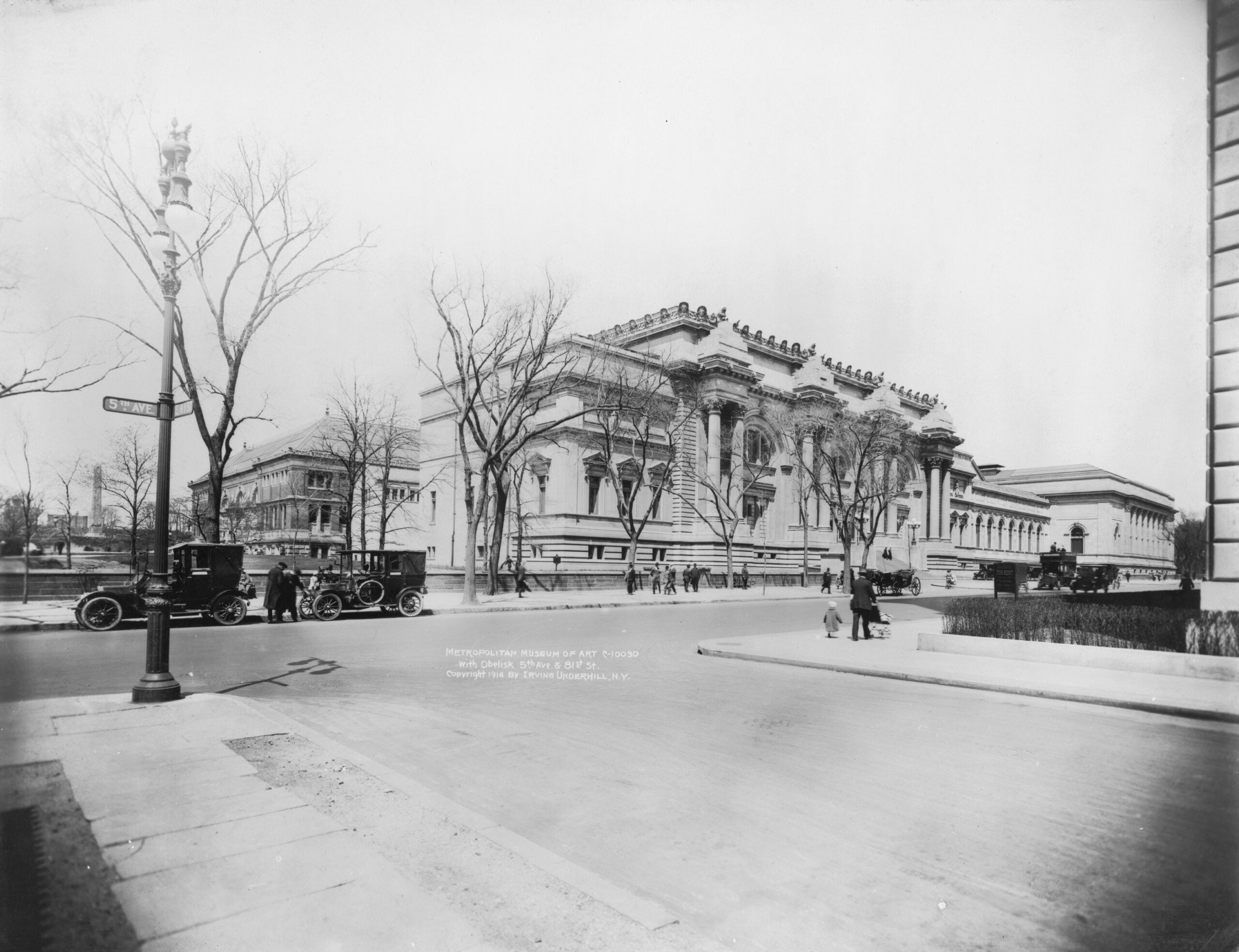 Black-and-white photo of a large neoclassical building with columns, people walking, leafless trees, and vintage cars parked by the sidewalk. A street sign is visible in the left foreground.