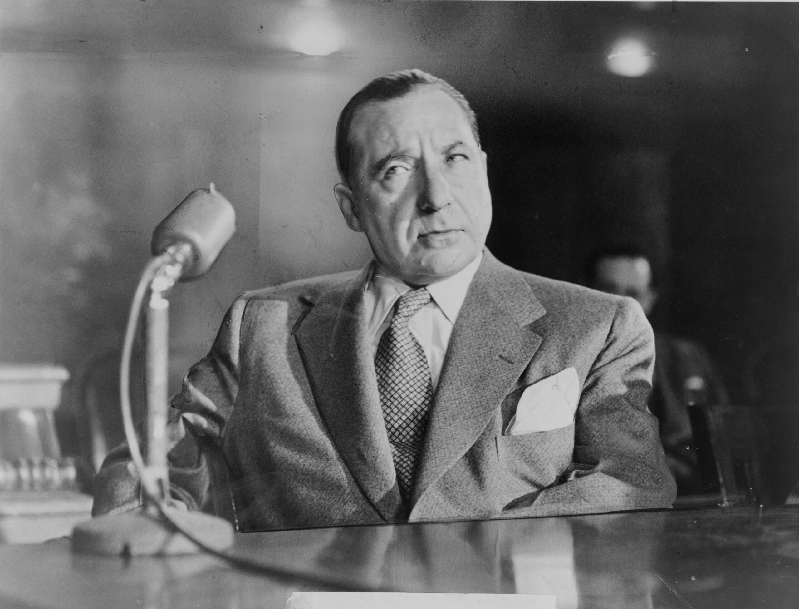 A man in a suit sits at a table with a microphone in front of him, looking to the side with a serious expression. The background is out of focus, giving a formal atmosphere.