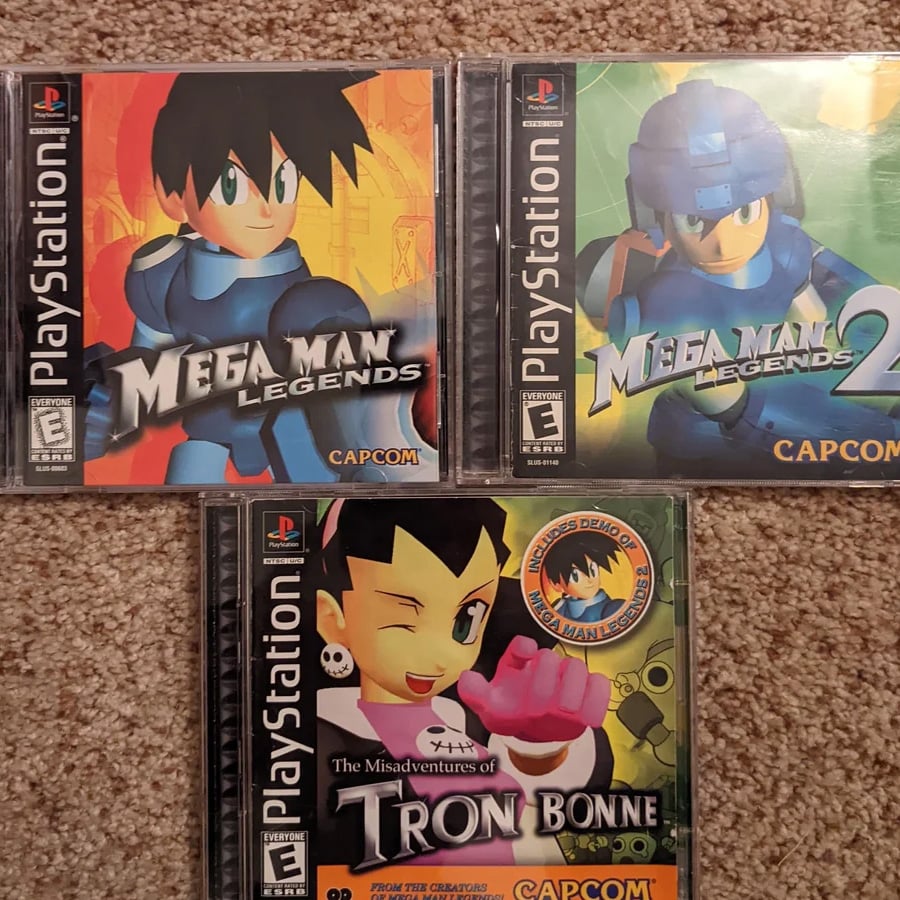 Three PlayStation game cases on a carpet: Mega Man Legends, Mega Man Legends 2, and The Misadventures of Tron Bonne, each featuring colorful animated cover art and the CAPCOM logo.
