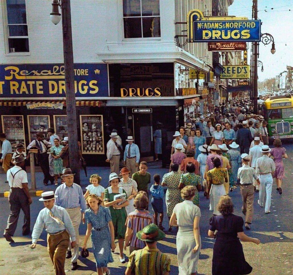 A busy street scene in the 1940s shows people crossing in colorful clothing. Drugstore signs, storefronts, and a green streetcar are visible. The crowd includes men, women, and children in summer attire.