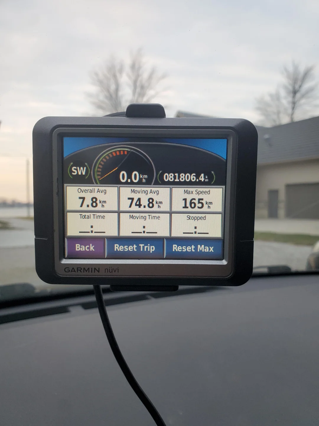 A Garmin nüvi GPS displays trip stats inside a parked car: 74.8 km moving avg, 7.8 km overall avg, 165 km max speed, 0.0 km/h current speed, 81,806.4 km total distance. House and trees visible outside.
