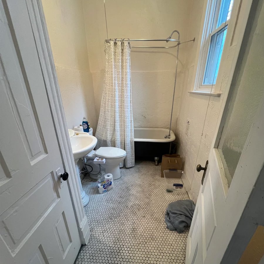 Small bathroom with hex tile floor, white sink, toilet, clawfoot tub with shower curtain, window, and some scattered items on the floor including a towel, box, and cleaning supplies.
