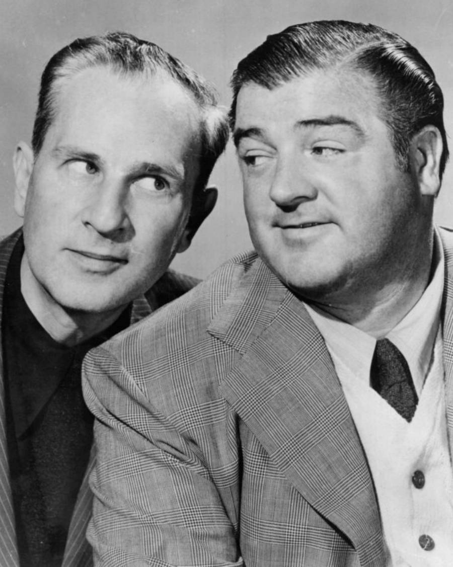 Two men in suits pose closely together, both looking to their right. One man has slicked-back dark hair and a checked jacket, while the other has lighter hair and a striped suit. The photo is black and white.