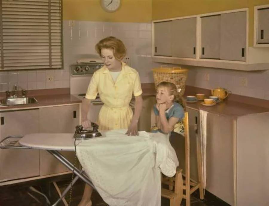 A woman in a yellow dress irons clothes in a retro kitchen while a young girl sits on a high chair nearby, watching her with interest. The kitchen has yellow cabinets and a basket of laundry in the background.