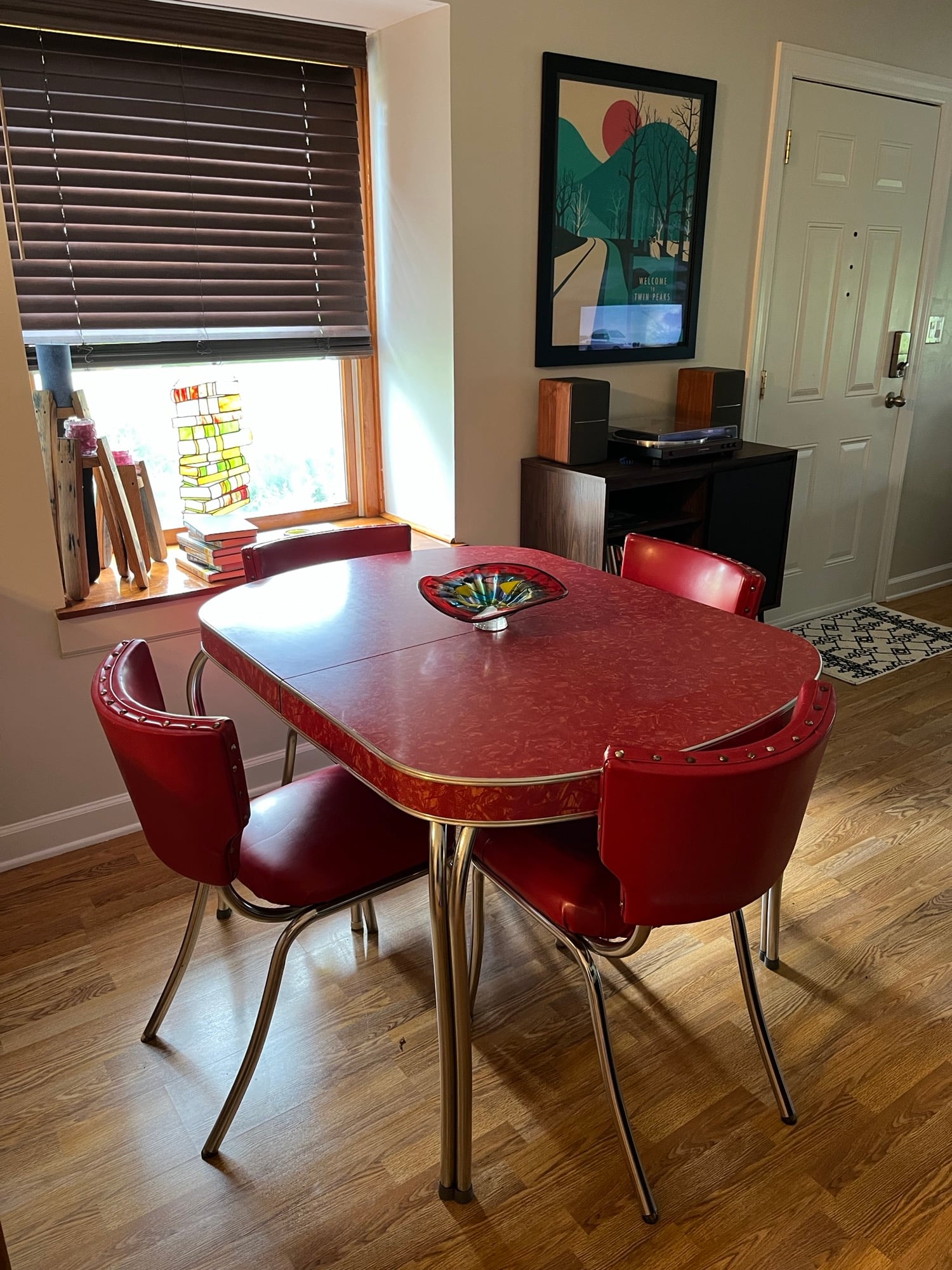A retro red dining table with four matching chairs sits on a wood floor near a window with blinds. A colorful dish is on the table. Wall art and a record player are visible in the background.