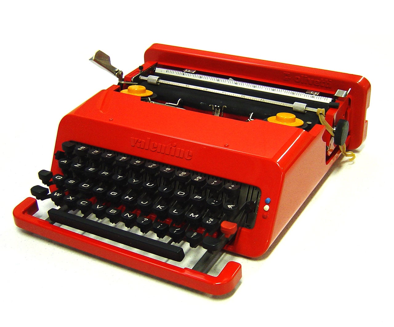 A bright red vintage typewriter with black keys and yellow knobs, set against a plain white background. The brand name “Valentine” is embossed on the front.