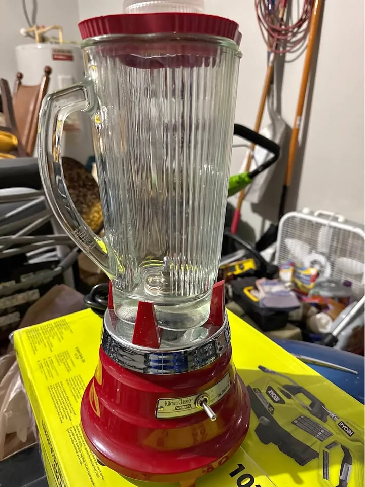 A vintage red and silver blender with a glass pitcher sits on a yellow cardboard box in a cluttered garage or storage area. Various household items and tools are visible in the background.