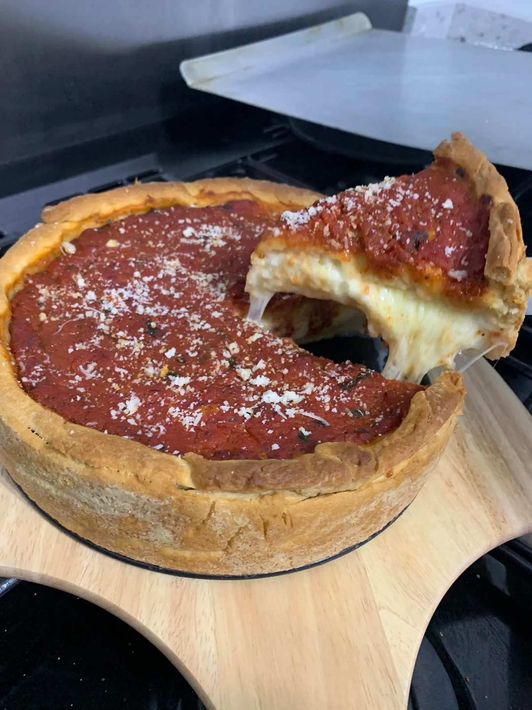 A deep-dish pizza with tomato sauce and cheese sits on a wooden board. One slice is being lifted, showing melted cheese stretching from the pizza.