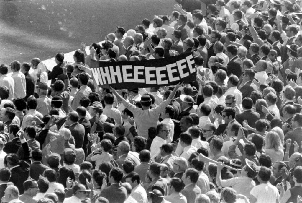 A person in a crowded stadium holds a large sign reading “WHHEEEEEE” as people around them watch an event.