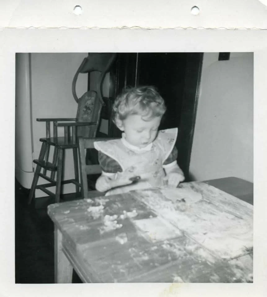 Black-and-white photo of a young child in a bib at a table, playing with dough or flour. A wooden high chair is seen in the background.