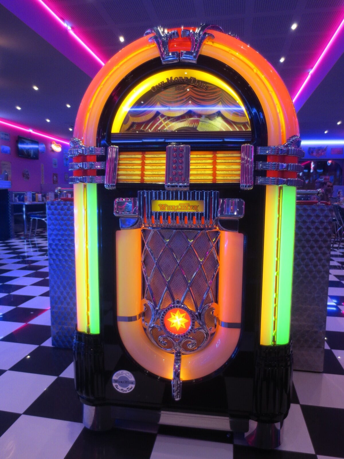 A brightly lit vintage jukebox with neon lights stands on a black-and-white checkered floor in a retro diner setting.