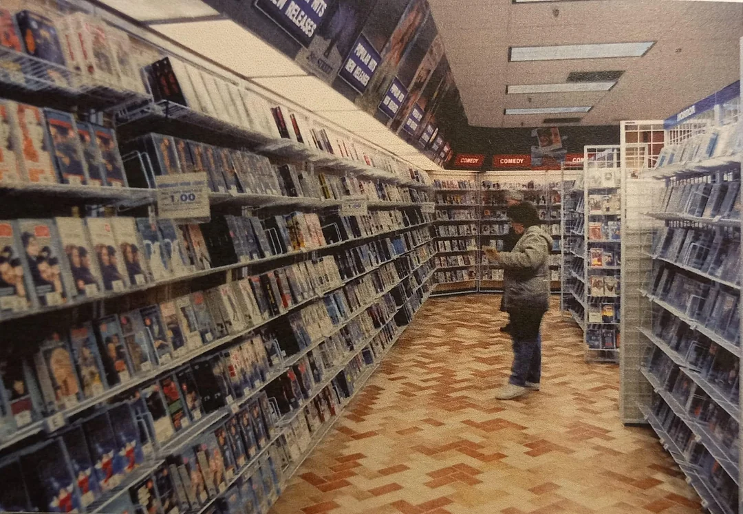 A person stands in an aisle of a video rental store, surrounded by shelves filled with DVD or VHS cases. The store has bright lighting and a checkered floor pattern.