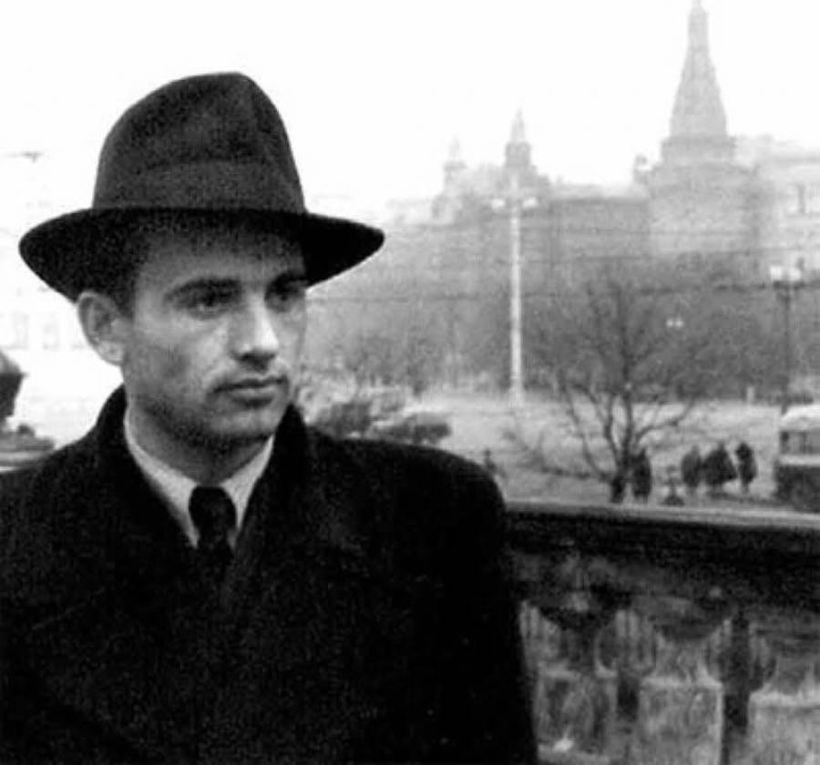 A man in a dark coat and hat stands outdoors, looking slightly to the side. The background is blurred, showing large buildings, bare trees, and a few people walking, giving a historical or vintage atmosphere.