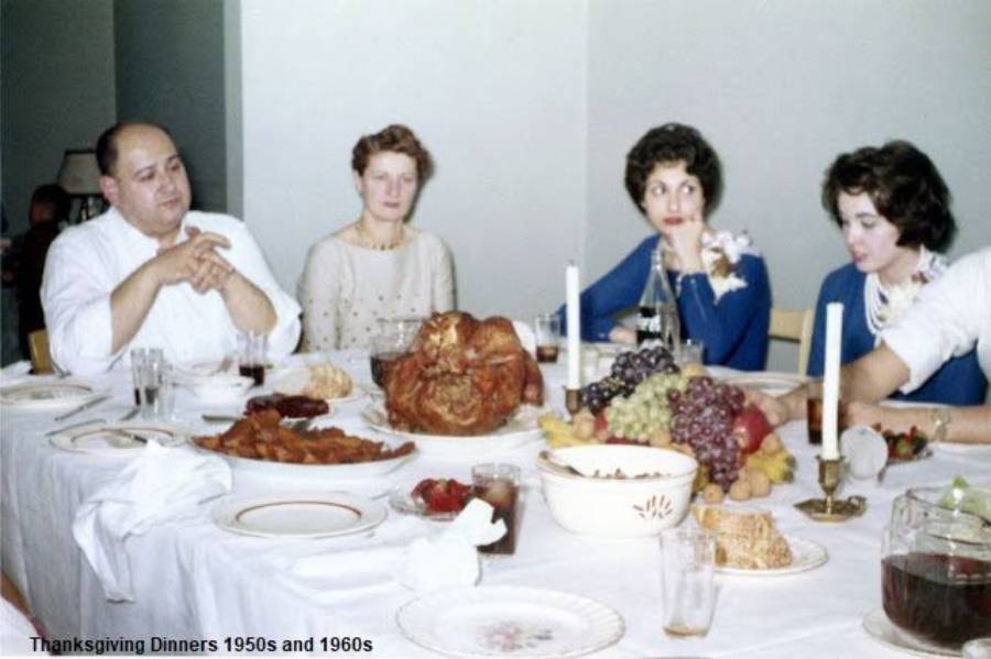 Four people sit around a Thanksgiving dinner table with a roast turkey, fruit, and various dishes. The setting appears to be from the 1950s or 1960s, with formal attire and candles on the table.