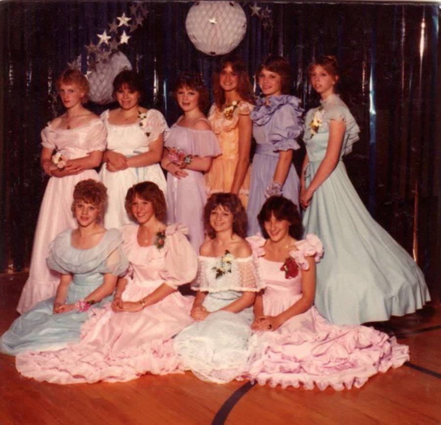 Nine young women in pastel, ruffled dresses pose in two rows at a formal event, such as a prom, with dark curtains, paper decorations, and star shapes in the background.
