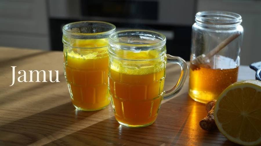 Two clear mugs of yellow jamu drink sit on a wooden table next to a jar of honey with a dipper and half a lemon. Sunlight streams in, and the word “Jamu” appears on the left.