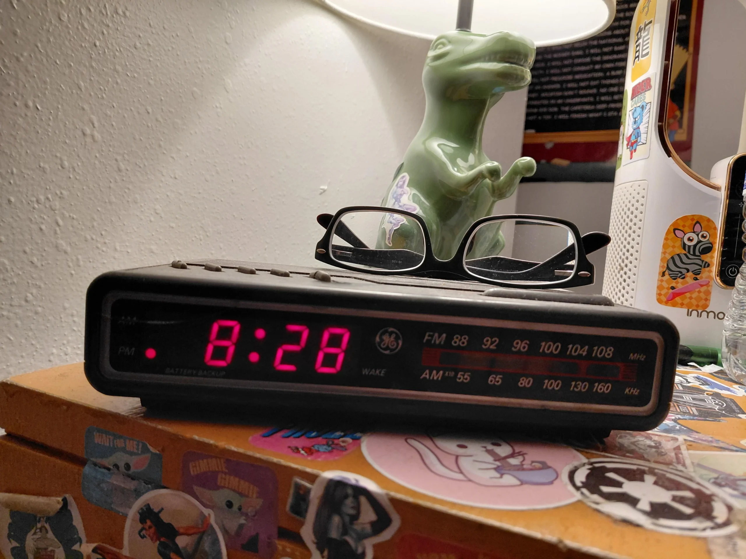A digital alarm clock showing 8:28 PM sits on a sticker-covered nightstand, with a pair of eyeglasses resting on top and a green dinosaur lamp in the background.