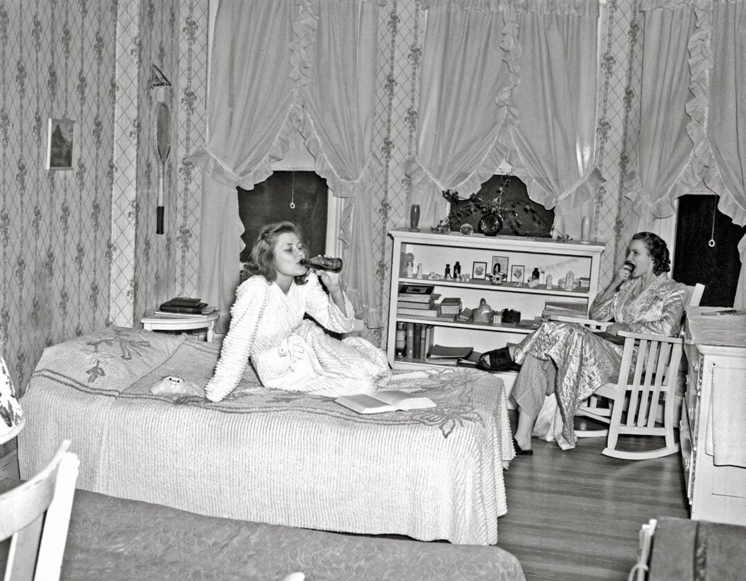 Two women in a vintage bedroom; one lounges on a bed drinking from a bottle, while the other sits in a rocking chair. Both wear robes in a room with patterned wallpaper, curtains, and shelves with books and photos.