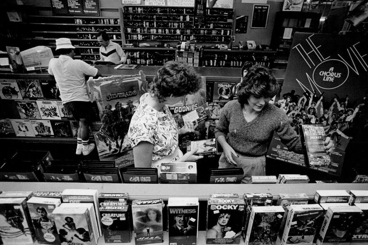 Black and white photo of people browsing vinyl records and VHS tapes in a crowded record store. Movie posters and album covers, including “The Goonies,” are visible. Two women look through records together.