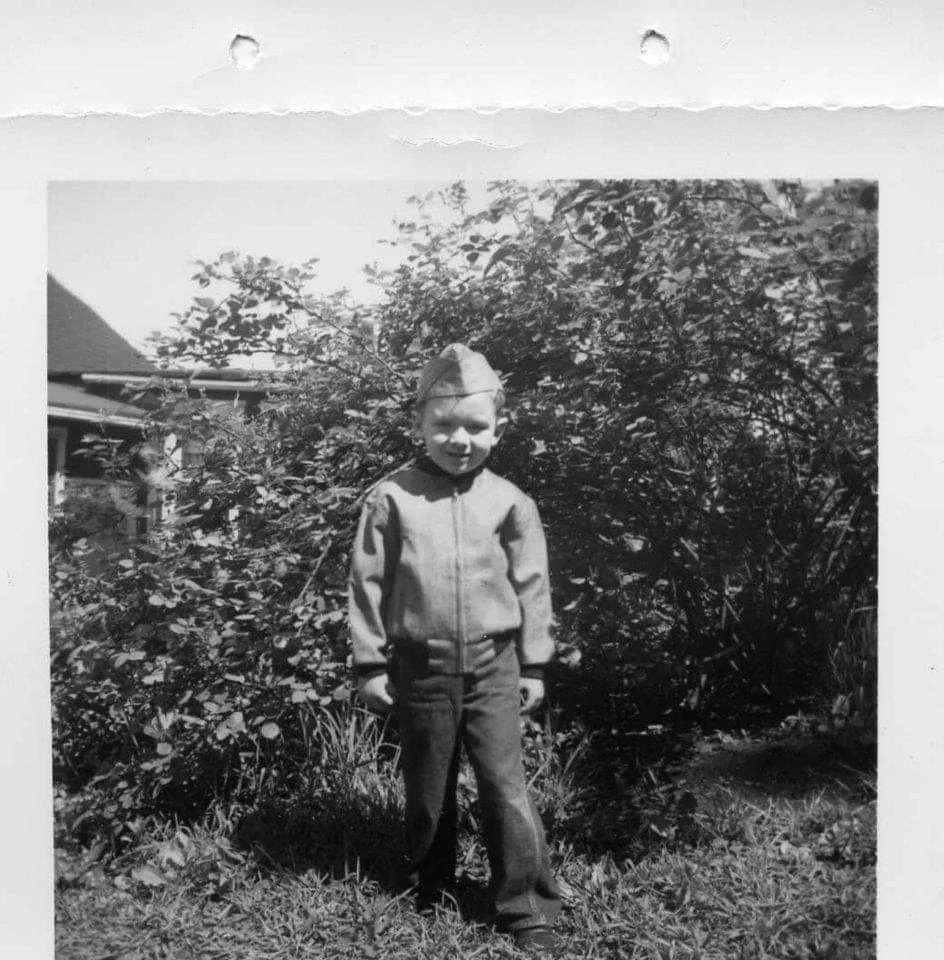 Black and white photo of a young boy standing outside in front of bushes, wearing a zippered jacket, pants, and a small pointed cap, and looking at the camera with a neutral expression.