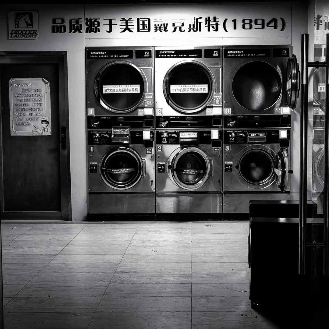 Black and white photo of a laundromat with six washing machines and dryers stacked in two rows. Signs with Chinese characters and numbers are visible above the machines. The floor and part of a bench appear in the foreground.