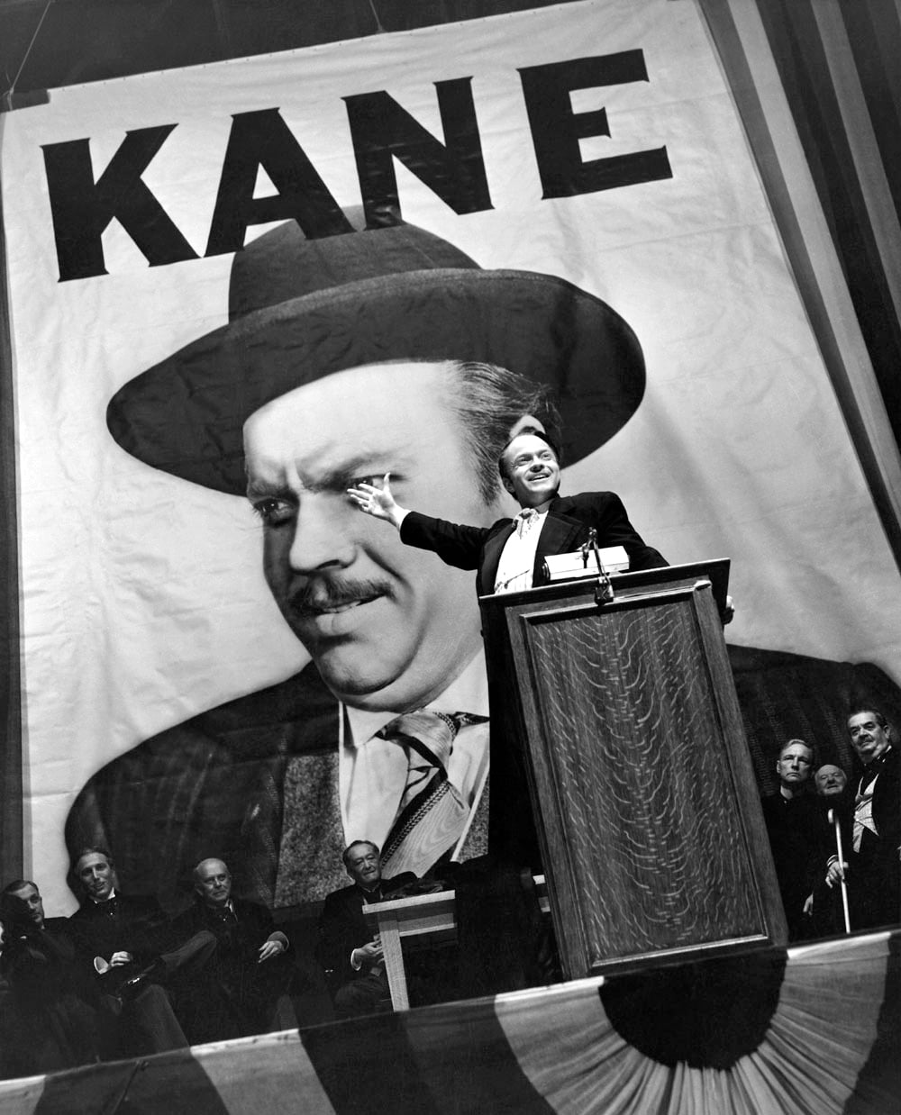 A man stands at a podium giving a speech, gesturing upward, with a large banner showing his face and the word “KANE” in bold letters behind him. Several people sit on stage in the background.