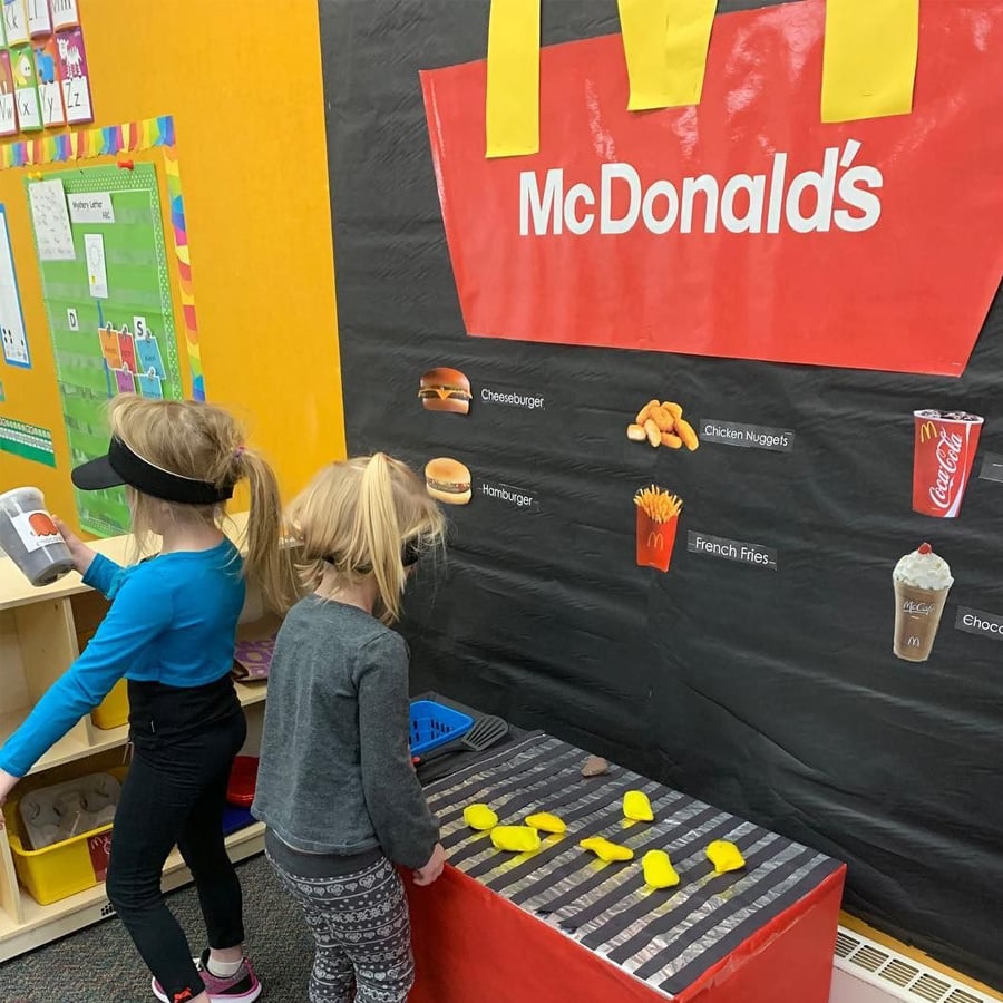 Two children play at a pretend McDonald’s stand in a classroom, grilling toy food and using props. The setup includes a black backdrop with a McDonald’s logo and menu items depicted on the wall.