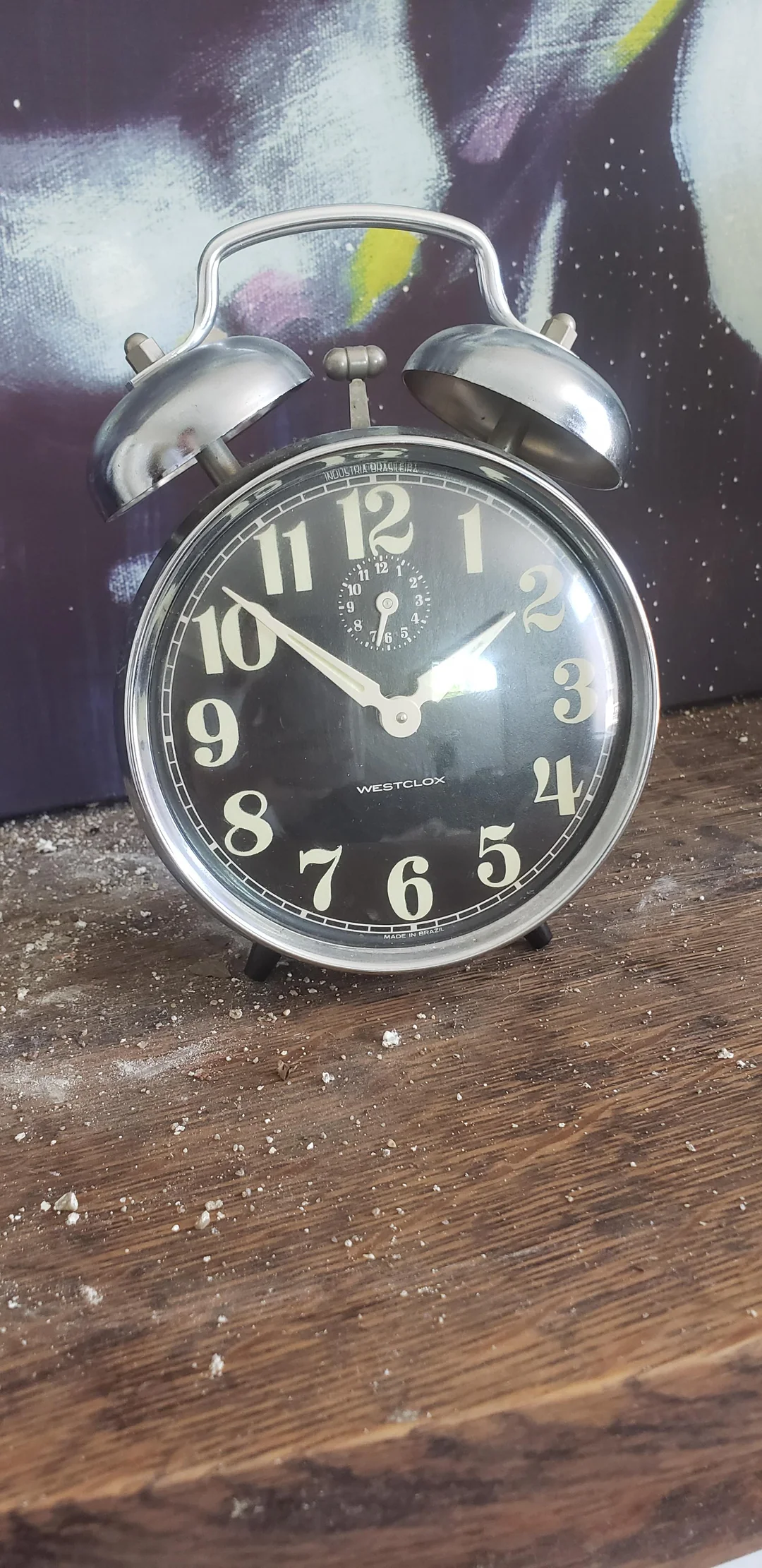 A vintage Westclox alarm clock with two bells sits on a dusty wooden surface in front of a painted background. The time on the clock reads 12:02.
