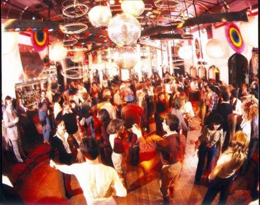 Crowded disco dance floor with people dancing under disco balls and colorful circular decorations, bathed in warm, vibrant lighting.