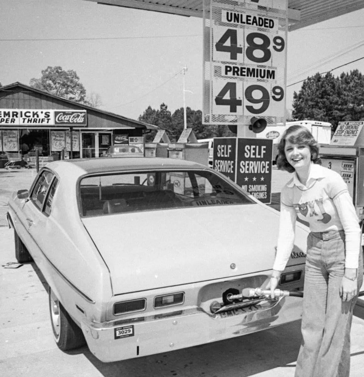 A woman smiles while pumping gas at a station. Gas prices are shown: unleaded 48.9 cents, premium 49.9 cents. A store and Coca-Cola sign are visible in the background.