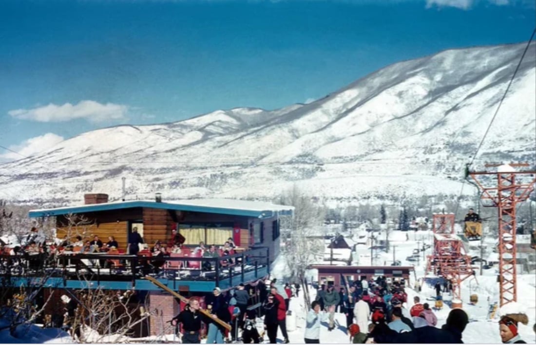 People gather at a ski resort with a snowy mountain backdrop, some on a balcony, others near a ski lift. The scene is lively under a bright blue sky.