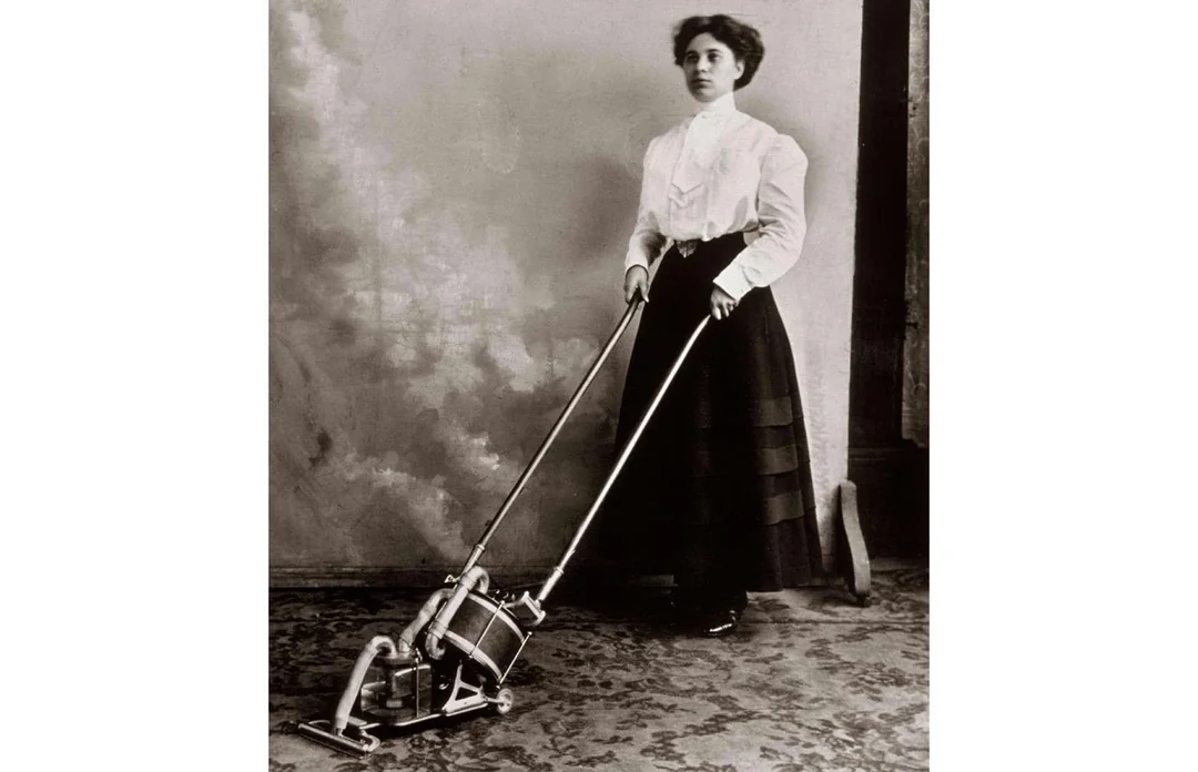 A woman in early 20th-century clothing operates an old-fashioned vacuum cleaner on a patterned carpet in a vintage, sepia-toned photograph.