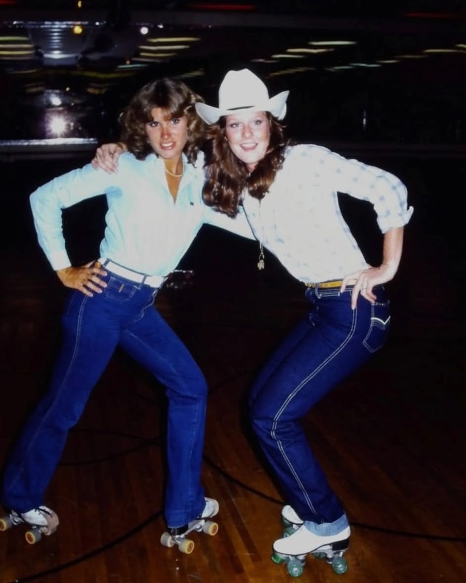 Two women in jeans and roller skates pose together indoors on a wooden rink. One wears a white cowboy hat and both have one arm on their hip, smiling at the camera under dim lights.